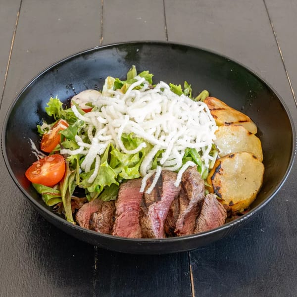 Steak salad with grilled potatoes, cherry tomatoes, lettuce, and crispy white noodles in a black bowl.
