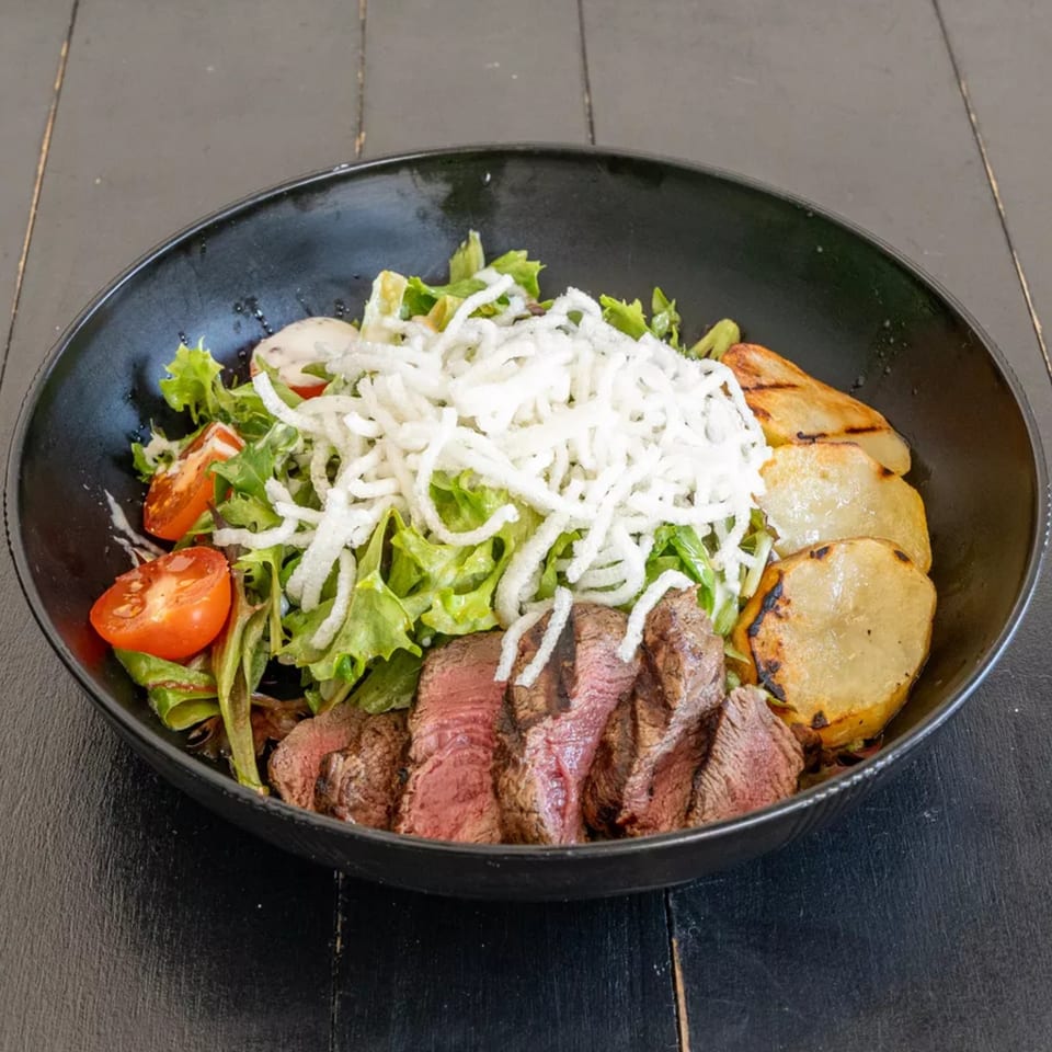 Steak salad with grilled potatoes, cherry tomatoes, lettuce, and crispy white noodles in a black bowl.