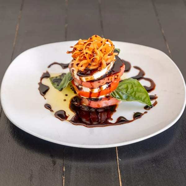 Stacked tomato and mozzarella with balsamic glaze, basil, and crispy fried onions on a white plate.