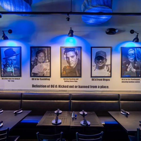 Kitchen 86 interior with black booths, tables, and celebrity portraits under blue lighting.